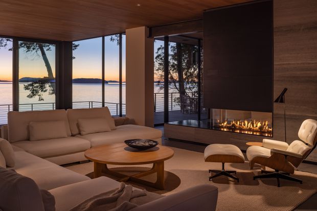 A refined contemporary living room at dusk with expansive floor-to-ceiling glazing overlooking a calm waterfront. The room features a large neutral sectional sofa, rounded timber coffee table, sculptural lounge chair and ottoman, warm timber ceiling, dark feature fireplace surround with a linear gas fire, and an open connection to an outdoor balcony or terrace. The atmosphere is warm, serene, and luxurious.
