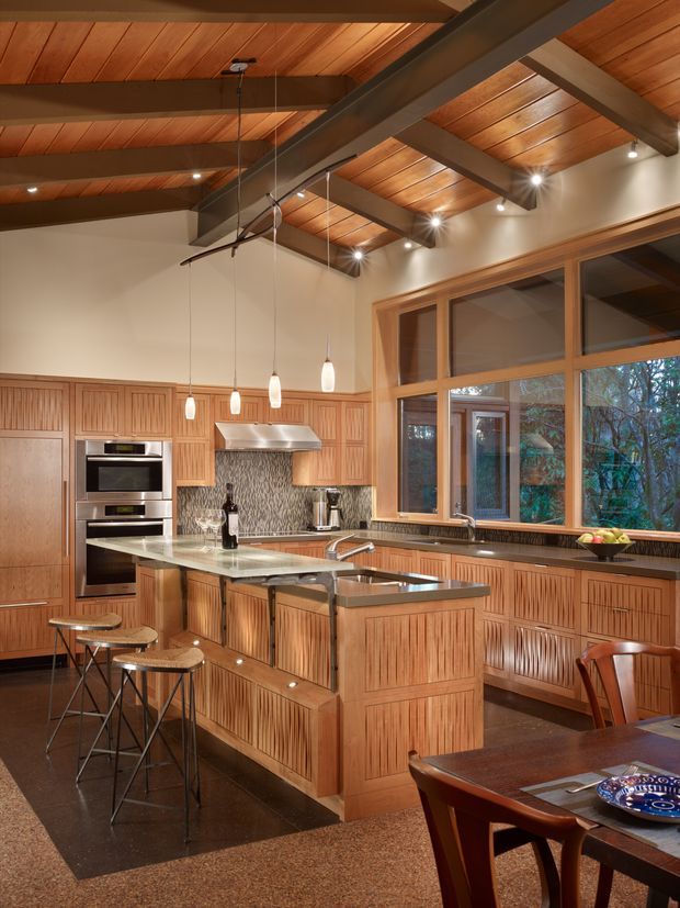Warm, generously proportioned kitchen and dining area with extensive custom timber cabinetry featuring vertical fluted and panelled fronts. A central island includes a pale stone benchtop, integrated sink and seating, while stainless-steel appliances and a textured patterned splashback sit along the rear wall. Exposed dark structural beams support a timber-lined vaulted ceiling with pendant and recessed lighting. Large timber-framed windows overlook trees, and a dining table occupies the foreground.