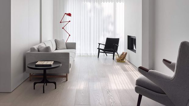 A bright, minimalist living room with pale timber flooring, white walls, sheer floor-to-ceiling curtains, and streamlined grey seating. A sculptural red wall lamp, black lounge chair, round black coffee table, and built-in fireplace add restrained graphic accents.
