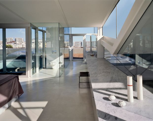 Bright contemporary open-plan interior with floor-to-ceiling glazing, polished concrete flooring, a long grey stone kitchen island or counter, and strong geometric architectural framing. A glass-enclosed bathroom area is visible at left, while an outdoor terrace and urban buildings can be seen beyond the glazing.