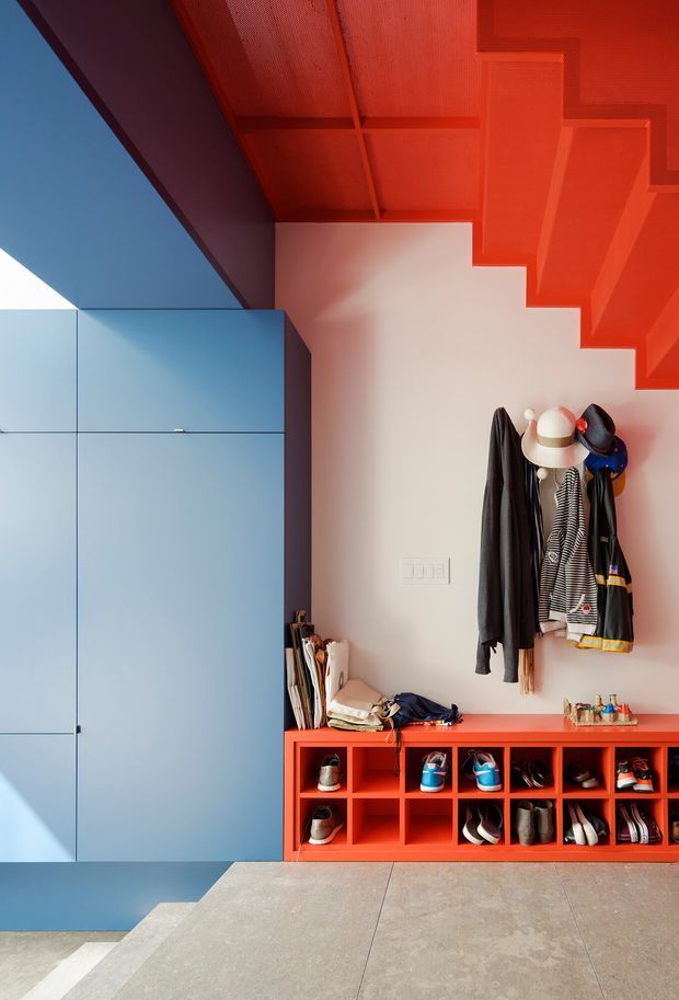Vibrant modern entryway with a tall pale-blue built-in storage cabinet, bright red overhead stair structure and matching red shoe cubbies, coats and hats on wall hooks, and large grey concrete-look floor tiles. The space uses bold colour blocking and clean geometric built-in joinery.