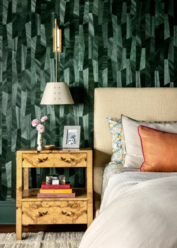 A richly layered bedroom vignette featuring a large upholstered bed with patterned and textured cushions, a warm burl-wood bedside table, brass wall-mounted reading light, floral arrangement, and framed photograph. The room is anchored by dramatic dark green patterned wallcovering and a neutral textured rug, creating an elegant, moody and eclectic atmosphere.