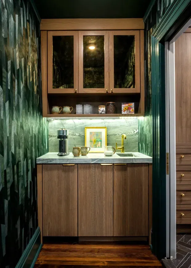 A compact, richly detailed beverage station or butler's pantry niche with fluted dark timber cabinetry, glass-front upper cupboards, a pale stone benchtop, integrated under-cabinet lighting, brass tapware and hardware, and a dark green patterned wall finish. Warm timber flooring and deep green trim create an intimate, moody, jewel-toned interior.