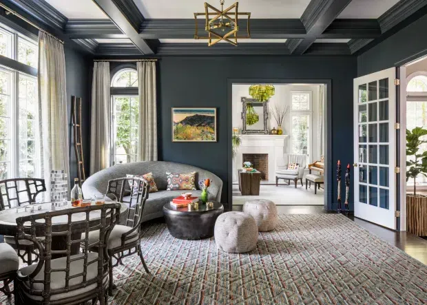 A richly layered formal living room with dark charcoal walls and coffered ceiling beams, tall multi-pane windows dressed in curtains, and a French-style glazed door opening to a bright adjoining sitting room. The room contains a curved sofa, woven dining or occasional chairs, a dark round coffee table, upholstered ottomans, a patterned area rug, a fireplace and traditional artwork. Brass geometric pendant lighting and dark architectural trim add a refined, heritage-inspired character.