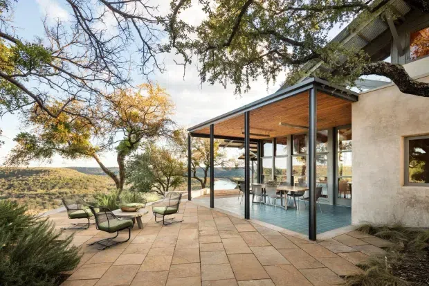 A contemporary hillside house with a broad paved terrace overlooking a wooded, semi-arid landscape. The terrace is furnished with low-profile outdoor lounge chairs and a fire bowl, while a glazed pavilion-like living or dining space extends beneath a flat roof with a warm timber-lined ceiling. Dark slender steel posts frame the extensive glazing, and pale rendered walls connect to the upper level of the home. Mature trees surround the setting.