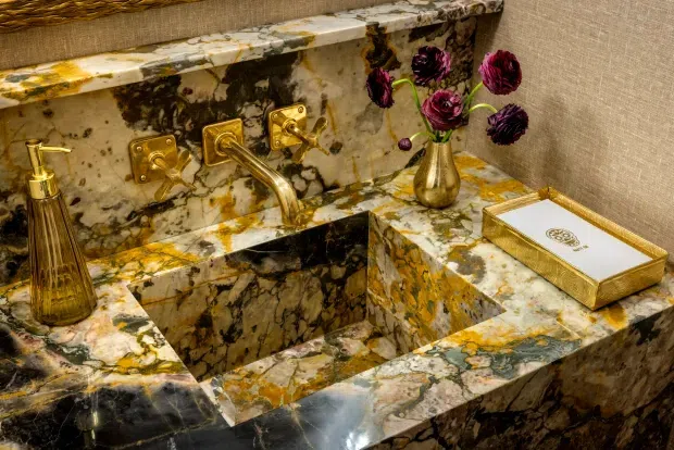 Luxurious bathroom vanity featuring a sculptural integrated stone basin and matching marble or onyx countertop and backsplash, with polished gold wall-mounted tapware and accessories. Dark burgundy flowers and a gold-trimmed tissue box provide decorative accents.