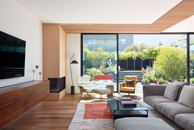Bright contemporary living room with warm timber flooring, a long dark timber media cabinet, white walls, and expansive black-framed glazing opening toward a planted courtyard. The room includes a grey sectional sofa, glass coffee table, lounge chair, floor lamp, and a linear timber slatted ceiling detail extending toward the glazing. The palette is warm and natural with strong indoor-outdoor connection.