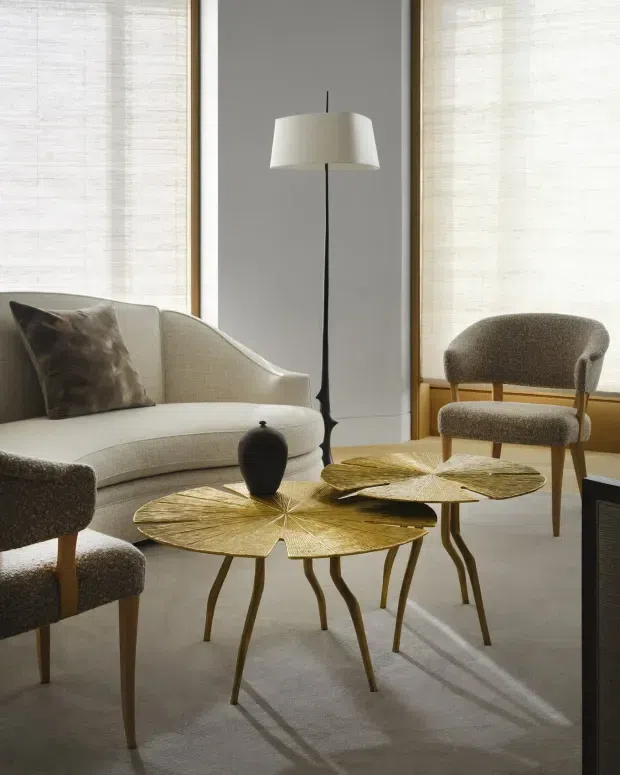 A refined living room with a cream upholstered sofa, matching textured lounge chairs, sculptural gold-toned leaf-shaped nesting coffee tables, a dark decorative vase, and a slim floor lamp. Large windows with light woven blinds provide soft natural light, while warm timber window framing and neutral finishes create an elegant, calm interior.