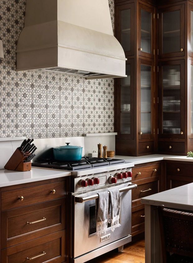 A warm, traditional kitchen centered on a stainless-steel range cooker beneath a large plaster range hood. Dark stained timber cabinetry with glass-front upper storage, a patterned tile backsplash, pale countertops, and timber flooring create a refined, heritage-inspired look.