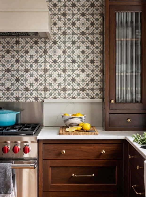 Detailed close-up of a traditionally styled kitchen featuring dark stained timber cabinetry with raised-panel fronts and brass hardware, white marble-look benchtops and splashback, a patterned geometric ceramic tile wall, and a stainless-steel gas range. A dark timber-framed reeded-glass cabinet adds vertical texture, while a bowl of lemons provides a bright accent.
