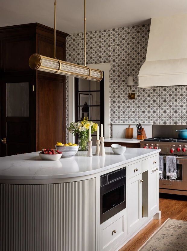 A refined kitchen featuring a large white island with fluted panel detailing, integrated oven, dark timber cabinetry, patterned geometric tile backsplash, white rangehood, brass pendant light, and a freestanding stainless-steel range. Warm timber flooring and classic hardware create a sophisticated, traditional-meets-contemporary interior.