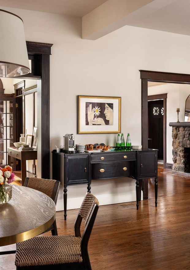 A refined dining and entertaining area with a round dining table and upholstered woven chairs in the foreground. A dark stained timber sideboard with brass-toned hardware sits against a light painted wall, topped with a coffee station and refreshments. Dark timber-trimmed openings connect to adjacent rooms, including a space with a stone fireplace. The room features polished timber flooring, framed artwork, traditional architectural trim, and warm layered lighting.