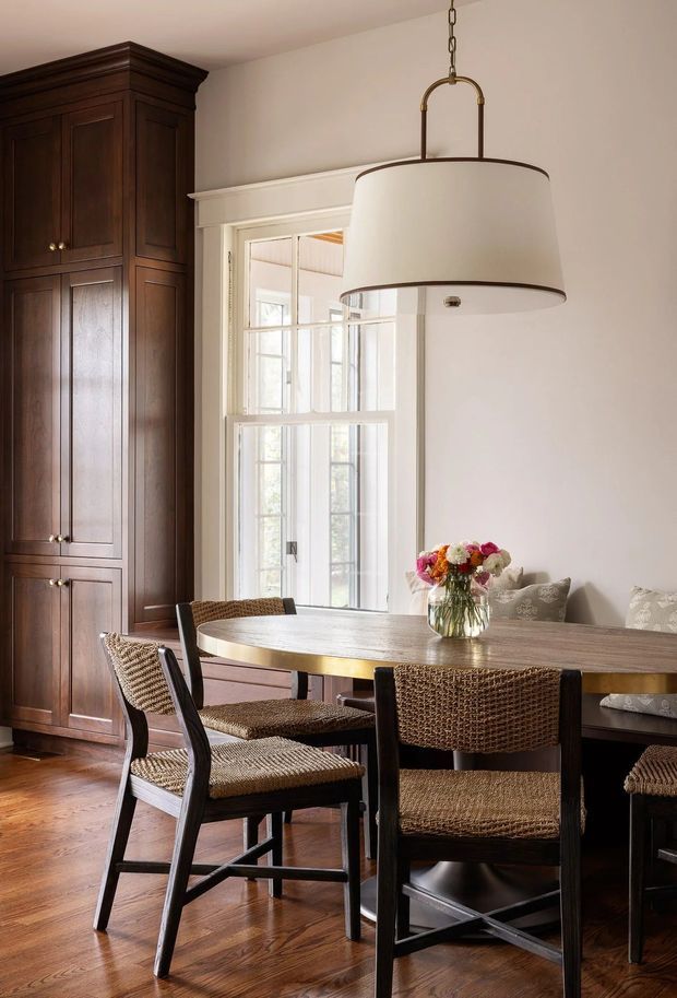 A warm, refined dining room featuring a dark wood built-in cabinet, a large wood dining table with woven chairs, white walls and trim, a multi-pane window, and an oversized pendant light. A floral arrangement and upholstered bench seating add softness to the space.