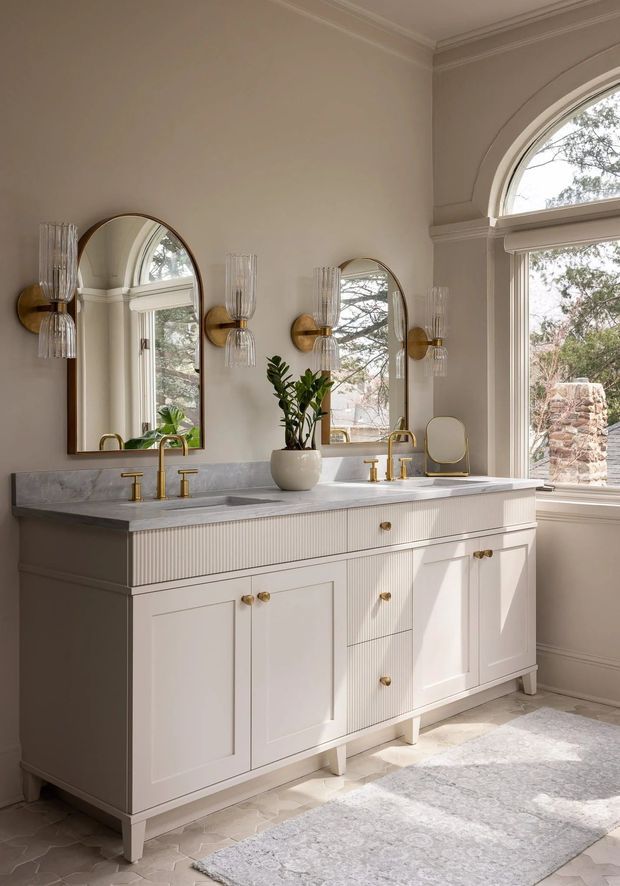 A refined traditional bathroom featuring a large double vanity with fluted white cabinetry, pale marble countertop, brass faucets and hardware, arched mirrors, decorative brass-and-glass wall sconces, and tall arched windows. Light patterned floor tiles and a soft neutral rug complete the warm, elegant interior.