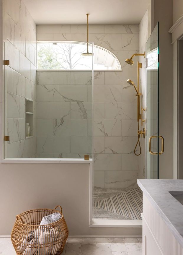 Bright, refined bathroom featuring a glass-enclosed walk-in shower with large white marble-look wall tiles, a curved clerestory window, brass shower fittings, a patterned mosaic shower floor, and a pale stone vanity. A woven basket adds a natural accent in the foreground.