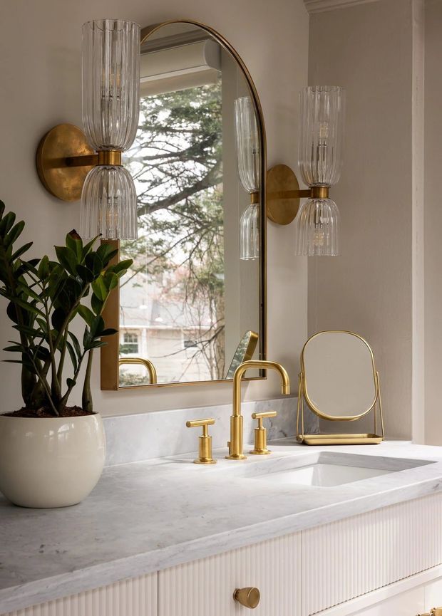 A refined bathroom vanity scene with a white marble countertop, integrated rectangular sink, fluted white cabinetry, and polished brass plumbing fixtures. A tall arched mirror with a brass frame reflects a window and tree outside, while matching fluted glass wall sconces flank the mirror. A potted plant and small brass-framed mirror add warmth and detail.