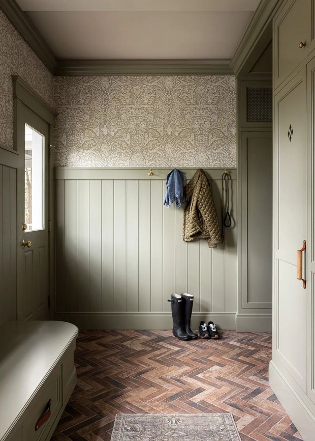 A refined mudroom or entryway with muted sage-green vertical wall panelling, patterned wallpaper above, ornate crown moulding, built-in cabinetry, hooks with coats, boots and shoes, and a dark herringbone brick or timber-look floor. A partially visible built-in bench sits along the left side, while a paneled door and tall storage cabinetry frame the room.