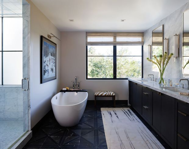 Spacious contemporary bathroom with a freestanding white bathtub, double vanity, large black-framed window, glass-enclosed shower, dark patterned floor tile, and a pale marble vanity top and runner-like floor inset. The room is bright and refined, with black cabinetry, large mirrors, and restrained decorative styling.