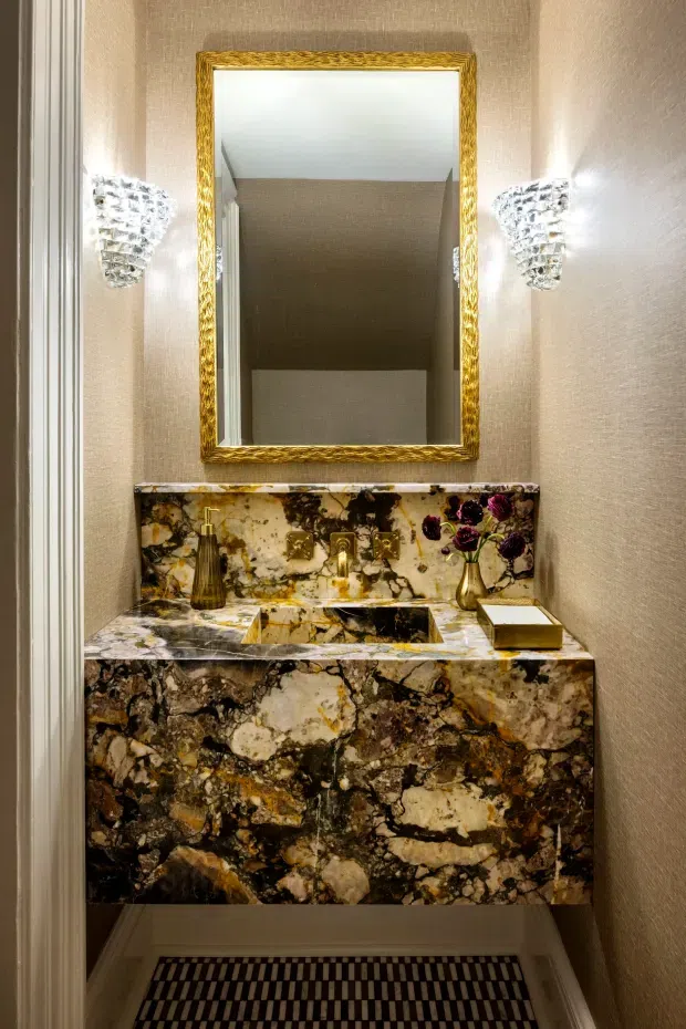 A compact, highly decorative powder room featuring a wall-mounted stone vanity with an integrated basin and dramatic veined dark marble or natural stone surfaces. A gold-framed mirror is flanked by crystal wall sconces, while textured neutral walls and ornate brass fixtures create a luxurious, glamorous interior.