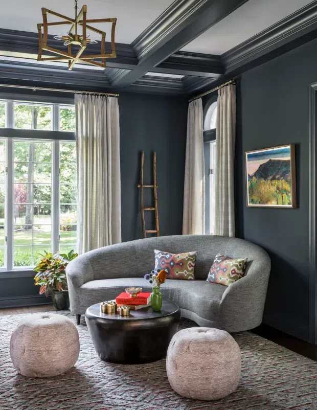 A refined living room with deep charcoal-blue walls, elaborate layered ceiling mouldings and a geometric brass-and-wood pendant light. Tall multi-pane windows with full-length curtains bring in garden views and daylight. A curved grey sofa, patterned cushions, rounded ottomans, a dark sculptural coffee table, artwork and a patterned rug create a sophisticated, eclectic seating area.