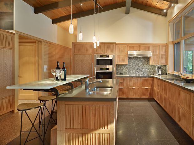 Spacious, warm-toned kitchen with extensive fluted timber cabinetry, a large central island with double sinks, integrated appliances, and bar seating. The kitchen features a dark stone-look benchtop, patterned mosaic splashback, polished dark flooring, pendant lights, and a timber-lined ceiling with exposed dark structural beams. Large glazed joinery on the right brings in natural light and views of greenery.