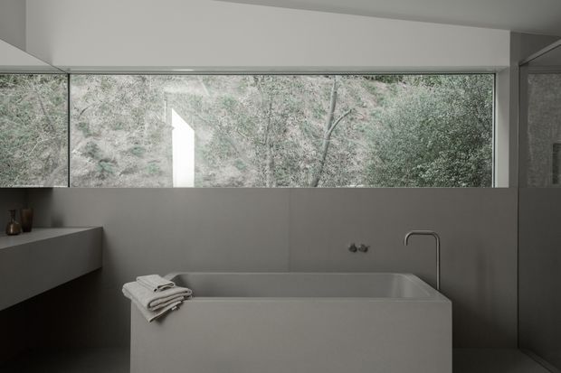 A minimalist contemporary bathroom with a freestanding rectangular bathtub positioned beneath a wide horizontal window overlooking dense greenery. The room features monochromatic grey finishes, a simple wall-mounted tap, a slim curved bath filler, integrated vanity joinery at left, and restrained natural styling.