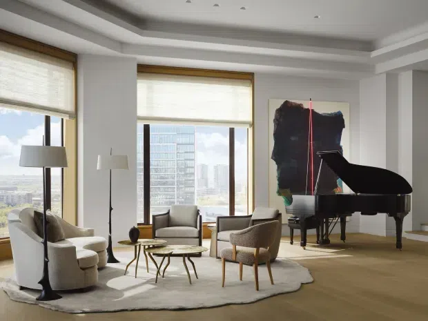 Elegant high-rise living room with expansive city-facing windows, layered recessed ceiling detailing, pale upholstered lounge chairs arranged around small sculptural coffee tables, a large light rug, and a black grand piano beside an abstract artwork. The space is bright, refined, and contemporary with warm timber flooring and light neutral finishes.