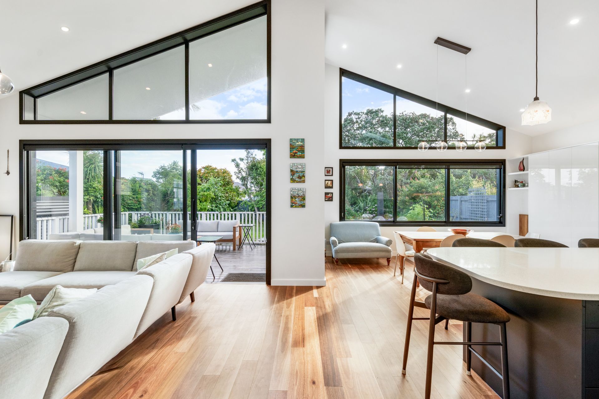 The full open-plan living, dining and kitchen at Devonport Bungalow — black steel glazing wrapping the space on both sides, timber floors running through, and the garden and deck never far away. Macfie Architecture.
