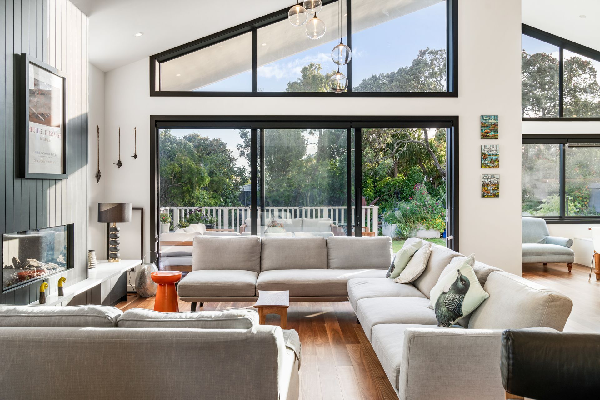 The living room at Devonport Bungalow — black steel stacking sliders and clerestory glazing dissolving the boundary between inside and garden, anchored by a dark shiplap fireplace wall and high stud ceiling. Macfie Architecture.