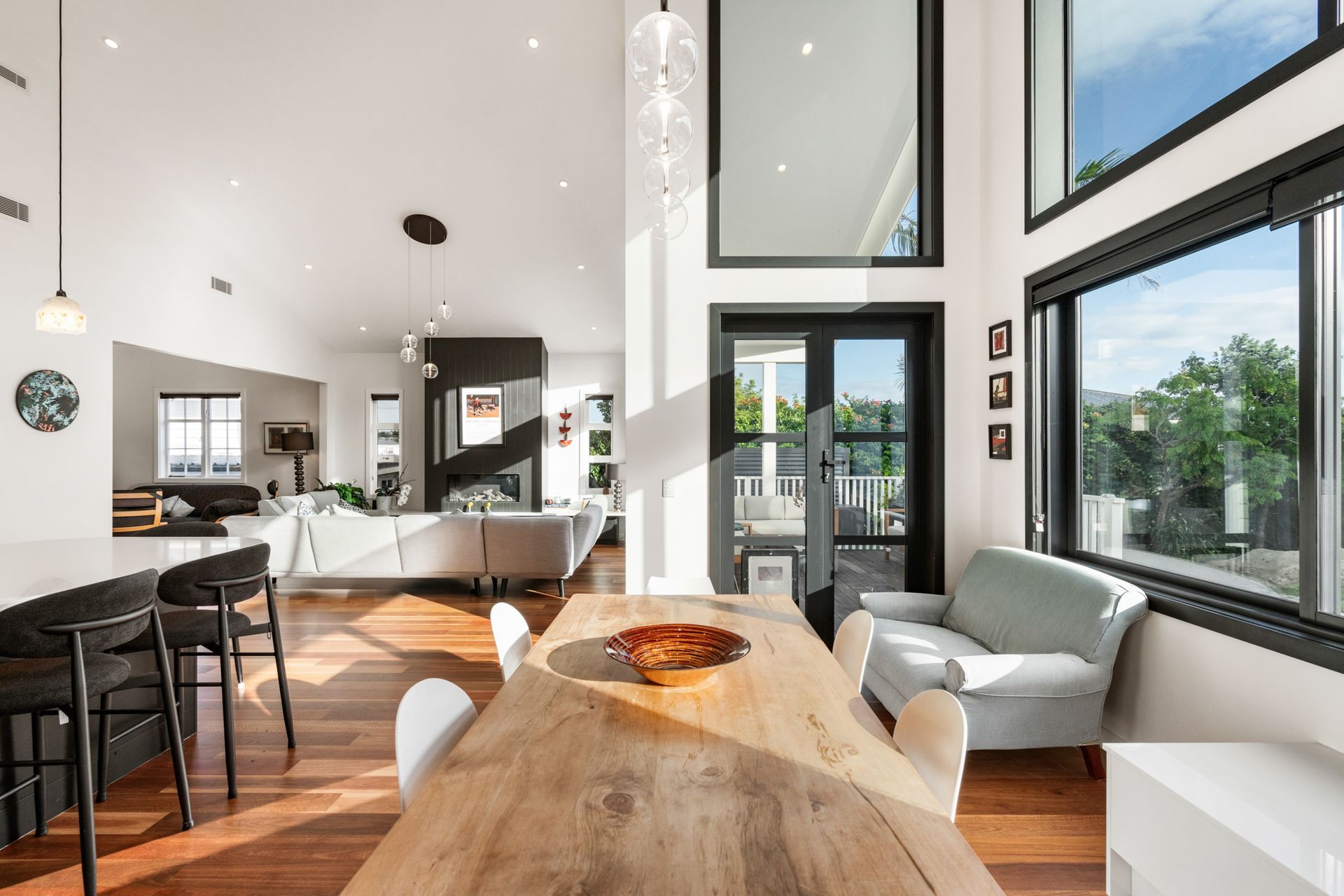 From island bench to dining table to fireplace — the full depth of the open-plan living at Devonport Bungalow, light flooding in through black steel joinery on every side. Macfie Architecture.
