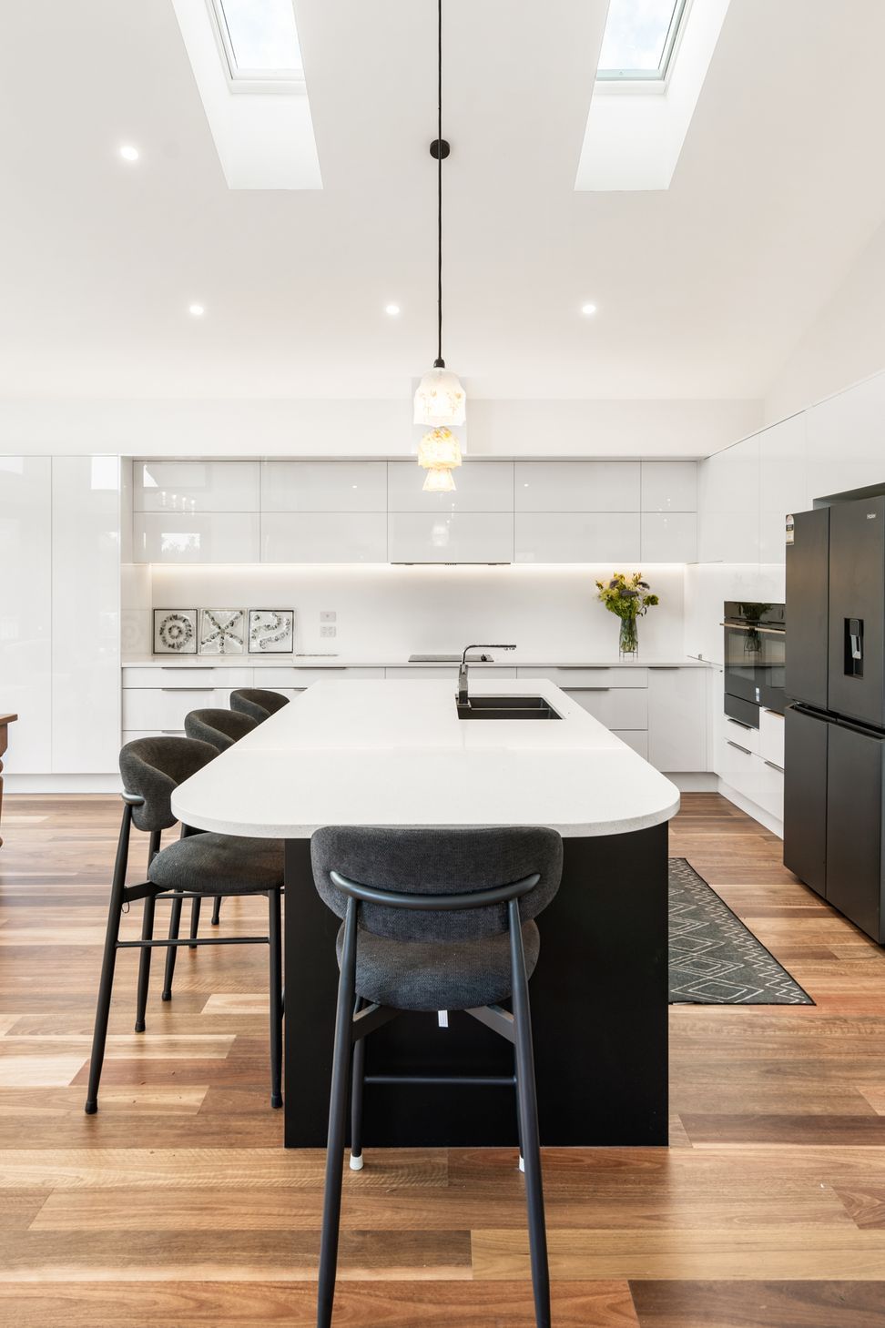 Skylights flooding the kitchen with natural light — gloss white cabinetry, dark island bench and white stone countertop at Devonport Bungalow. Macfie Architecture.