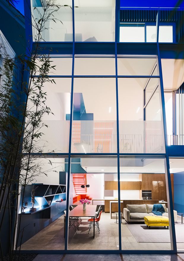 A tall, multi-level contemporary interior viewed through a blue-painted steel-framed glazed facade. The lower level contains an open-plan living and dining area with a grey sofa, bright yellow ottoman, dining table and chairs, dark built-in media joinery, and a warm timber kitchen. White walls, translucent ribbed glass panels, vivid red and blue accents, and strong geometric grid lines create a colourful, modern architectural composition. A narrow planted courtyard is visible along the left side.