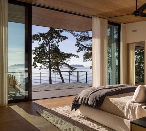 A warm, modern bedroom opening onto a waterfront deck through expansive black-framed glazed doors. The room features a timber-lined ceiling, timber flooring, full-height cream curtains, a large upholstered bed or daybed with neutral bedding, and a textured area rug. Calm water, trees, and a distant shoreline are visible beyond the deck.