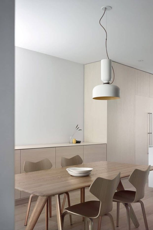 A minimalist open-plan dining area with a light timber dining table and sculptural wooden chairs. Pale timber built-in cabinetry lines the rear wall and continues into a tall kitchen storage bank, while a simple white pendant light with a warm metallic underside hangs above the table. The space has a restrained neutral palette, clean lines, and understated Scandinavian-inspired detailing.