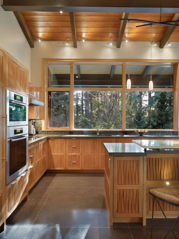 Warm, wood-lined kitchen with extensive custom cabinetry, a large central island, stainless-steel benchtops, integrated ovens, pendant lighting and a broad bank of timber-framed windows overlooking dense trees. Exposed timber ceiling boards and substantial beams add a strong natural architectural character.
