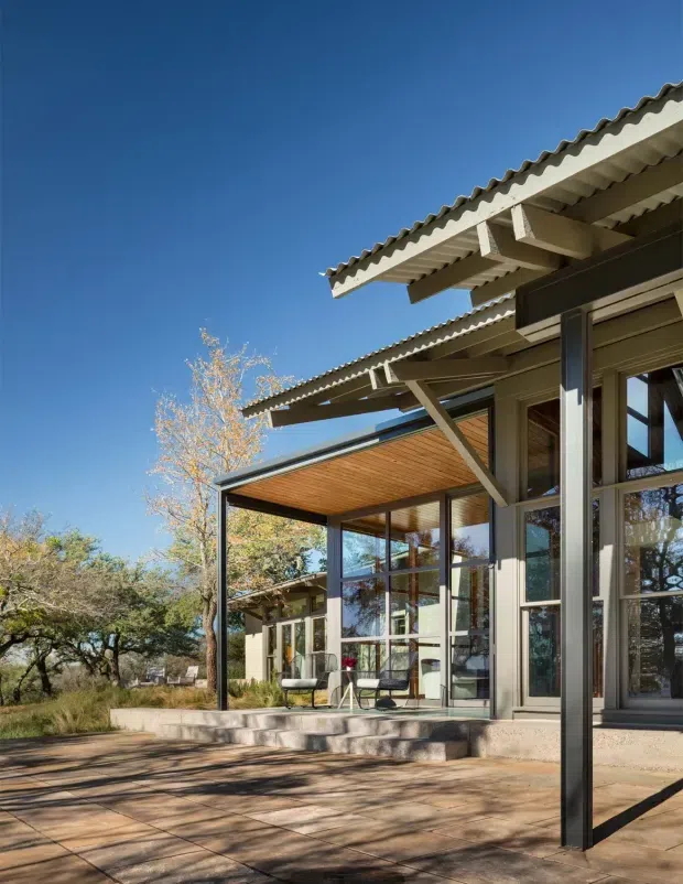 Exterior view of a contemporary single-storey building opening onto a broad paved terrace. The facade combines extensive floor-to-ceiling glazing with dark metal framing, vertical timber-toned cladding, and projecting corrugated roof planes supported by exposed timber rafters and angled braces. A covered outdoor sitting area is visible, surrounded by mature landscape under a clear blue sky.