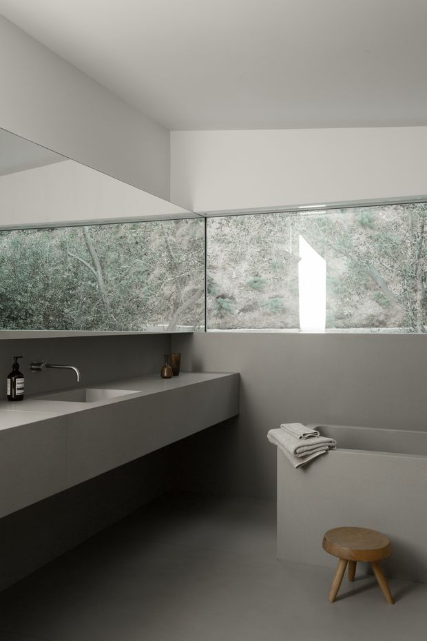 A minimalist contemporary bathroom with a floating integrated vanity and rectangular basin on the left, wall-mounted brushed metal tapware, a freestanding bathtub on the right, and a large horizontal window overlooking trees. The room has smooth pale grey surfaces, dark grey flooring, frameless glazing, and restrained natural timber accents from a small stool. Soft daylight creates a calm, monochromatic atmosphere.