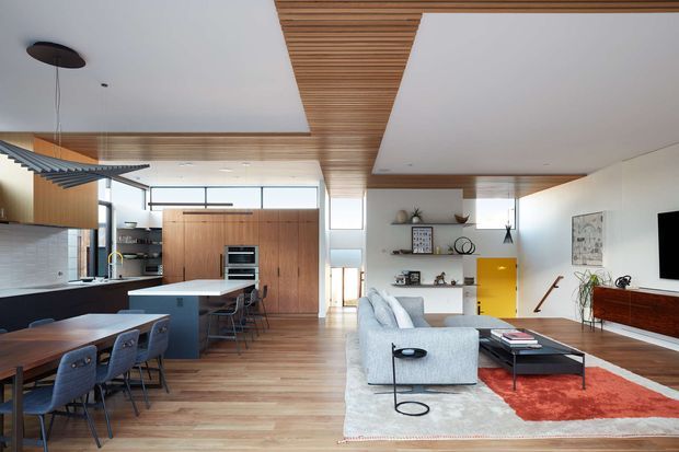Open-plan contemporary home interior combining a kitchen, dining area, and living room. The space features warm timber flooring and timber ceiling detailing, white walls, extensive glazing, a dark kitchen island with an integrated dining table, minimalist shelving, a pale sectional sofa, and a vivid red area rug. Clean-lined mid-century-inspired furniture and colourful accents create a bright, refined atmosphere.