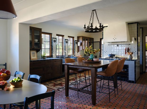 Open-plan kitchen and dining area with dark timber cabinetry, a large central island with seating, a separate dining table, tall timber-framed windows, patterned terracotta floor tiles, and a tiled cooking alcove with a white range hood. A traditional wrought-iron chandelier hangs over the dining table, while colourful patterned surfaces and warm timber create an eclectic, characterful interior.