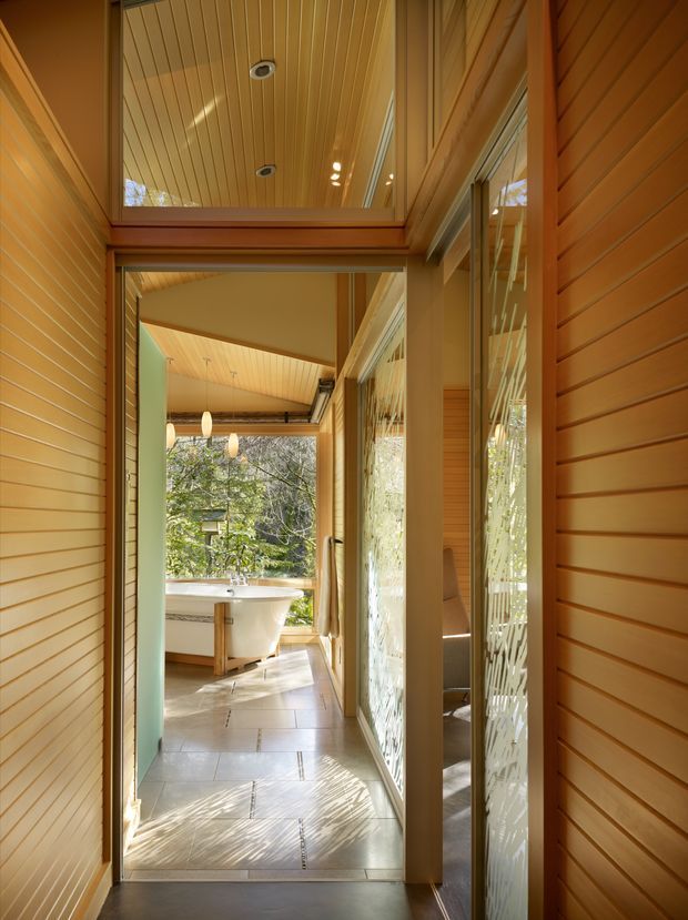 A warmly lit timber-lined hallway leading directly into a bathroom, with a freestanding white bathtub visible at the far end. The corridor features extensive horizontal timber wall panelling, a timber-lined ceiling with recessed lights, glazed openings and reflective surfaces, light tiled flooring, and strong natural light filtered through nearby trees. The composition highlights a calm, connected transition between circulation space and bathing area.
