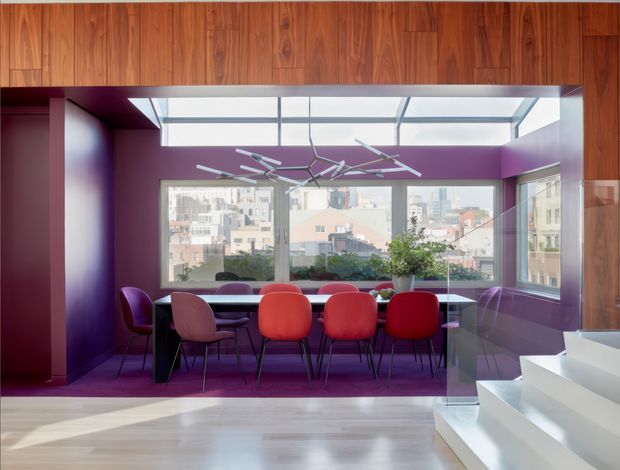 A vibrant contemporary dining area with a long dark dining table and colourful upholstered chairs, enclosed by floor-to-ceiling windows overlooking an urban cityscape. Deep purple walls, a timber-lined ceiling and upper wall band, a sculptural suspended light fitting, and a white stairway with glass balustrade create a bold, polished interior.