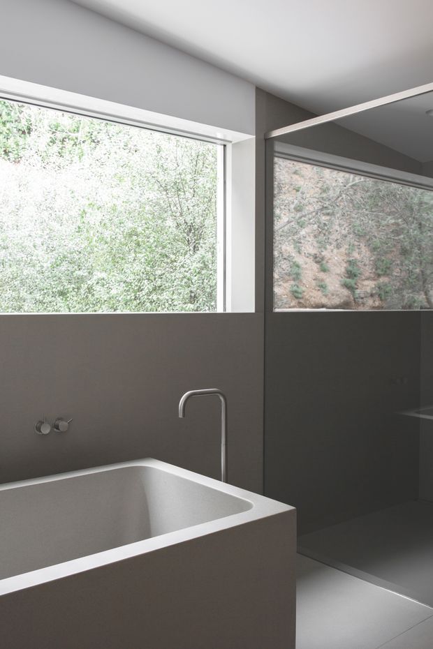 Minimalist contemporary bathroom with a sculptural freestanding rectangular bathtub, matching wall-mounted bath controls, a slim curved metal tap, and a frameless glass shower enclosure. Dark grey smooth wall surfaces contrast with white ceiling and window frames, while large windows bring in abundant natural light and overlook greenery and a rocky slope.