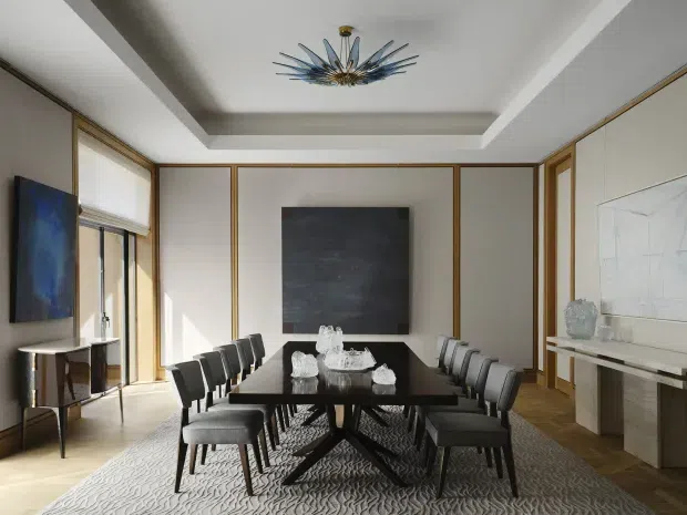 A refined dining room with a long dark dining table and upholstered chairs, framed wall panelling, timber flooring, a large textured rug, and a sculptural blue-and-gold pendant light set within a recessed ceiling. A minimalist console, artwork, and fireplace or display ledge are visible around the room. The palette is neutral with warm timber and metallic detailing.