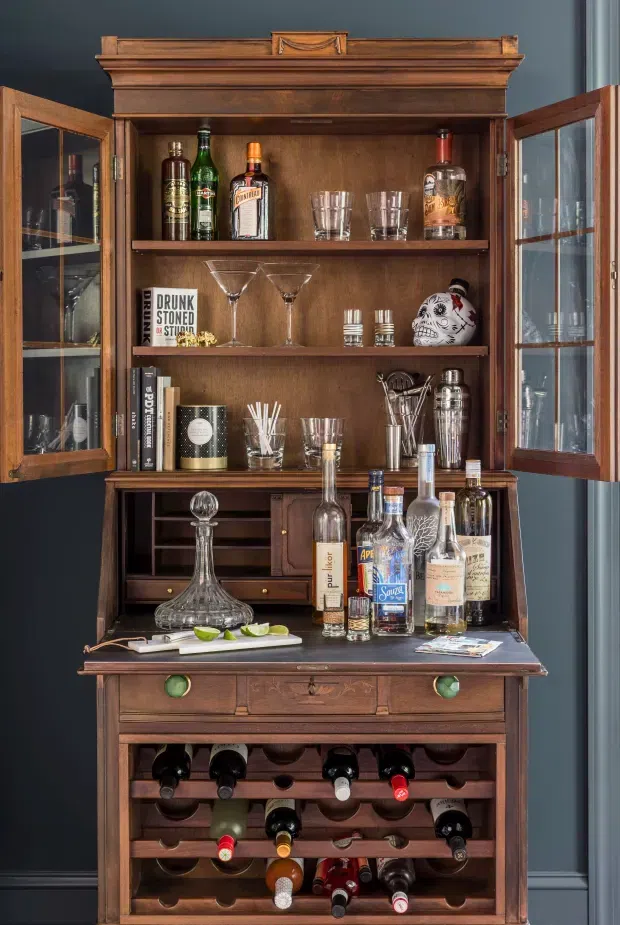 A compact, richly detailed home bar housed in a traditional dark timber drinks cabinet. The cabinet has glazed swing doors, open shelving with spirits and glassware, a lower preparation counter with bottles and cocktail tools, and integrated wine storage drawers and racks. It is set against a deep blue-grey wall and styled with warm wood, brass-toned hardware, and vintage bar accessories.