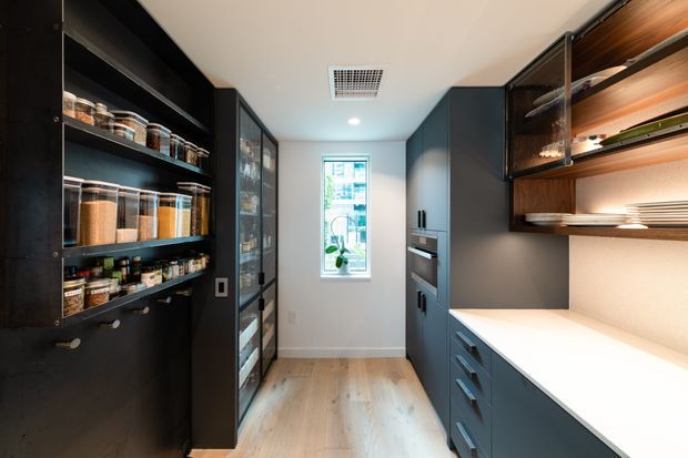 A narrow, compact walk-in pantry with dark blue-grey built-in cabinetry, open shelving filled with pantry goods, a bright white benchtop, integrated appliances, and warm timber shelving accents. Light timber flooring and a small window brighten the otherwise refined, contemporary space.