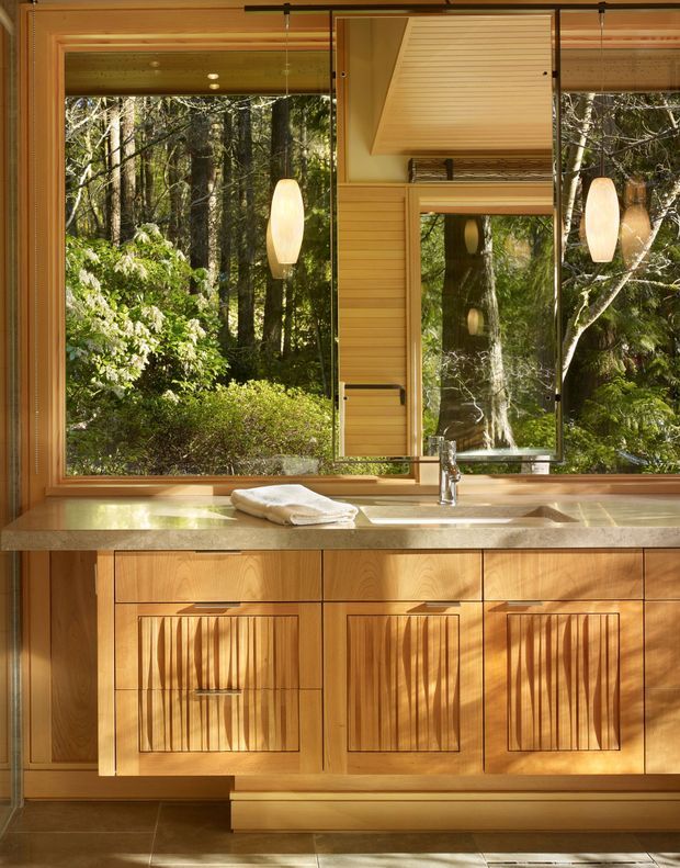 A warm, compact kitchen or kitchenette focused on a timber-framed window overlooking dense forest. The room features extensive honey-toned timber cabinetry with distinctive vertical slatted door fronts, a pale stone benchtop with an integrated sink and chrome tap, timber wall panelling, and a large operable window with reflective glazing. Natural light and woodland views create an organic, calm atmosphere.