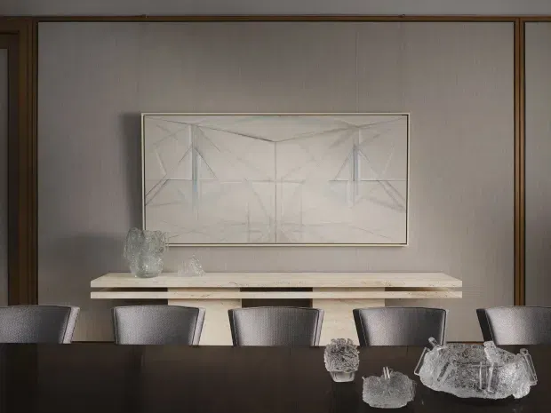 A refined dining room viewed across a dark dining table, with upholstered dining chairs, a long pale timber console, textured neutral wall panelling, dark timber framing, and a large minimalist geometric artwork. Clear glass decorative objects form a subtle centrepiece on the table. The atmosphere is understated, warm, and sophisticated.