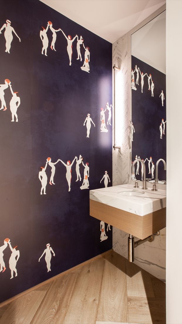 A compact, highly stylised bathroom or powder room with a floating timber vanity, white marble countertop and integrated basin, chrome tapware, and a large mirrored wall. The dominant navy wall covering features repeated white fashion-figure silhouettes with red accents. Light timber flooring and pale textured wall tile add contrast, while exposed chrome plumbing is visible below the vanity.