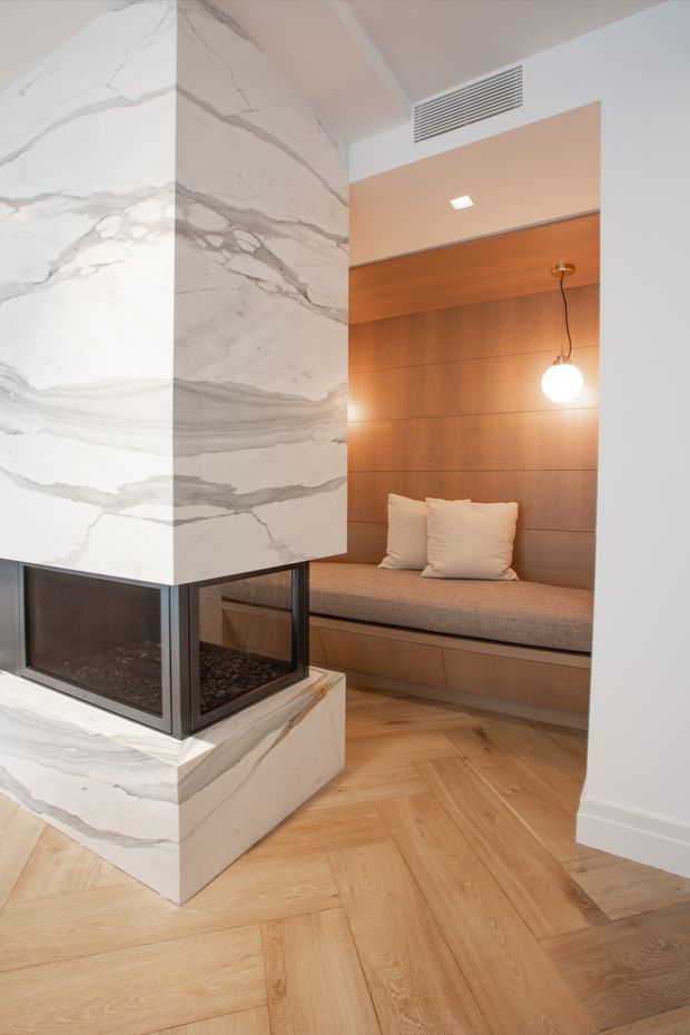 A refined contemporary interior featuring a double-sided or freestanding fireplace clad in dramatic white veined stone, positioned beside a built-in timber reading nook. The nook has warm wood wall panelling, integrated bench seating with upholstered cushion, two white pillows, and a suspended spherical pendant light. The floor is laid in a natural timber herringbone pattern, with white walls and a linear ceiling air vent.