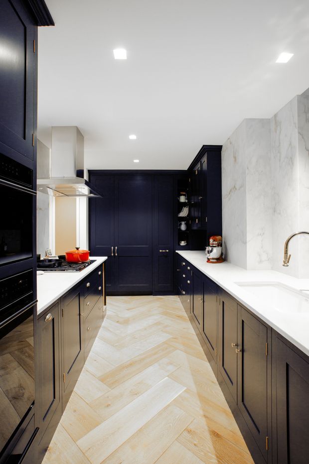 A narrow, refined galley kitchen with dark navy-black panelled cabinetry on both sides, white stone benchtops, a large veined white stone splashback, integrated dark appliances, brass hardware and tapware, and a pale timber herringbone floor. Recessed ceiling lights provide bright, even illumination, while a rear door and open shelving complete the compact layout.